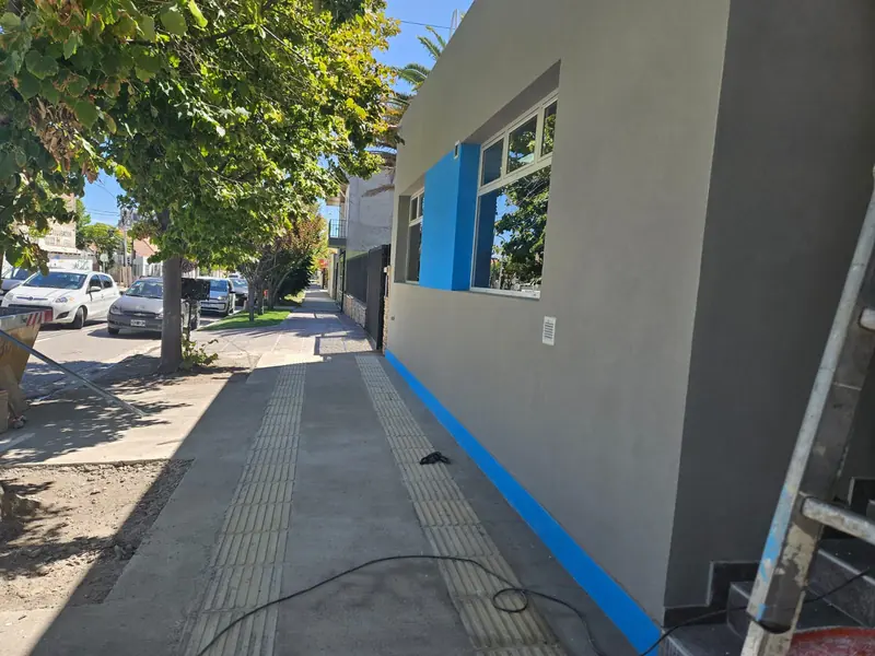 Vereda lateral terminada junto a la fachada gris con franja azul del colegio