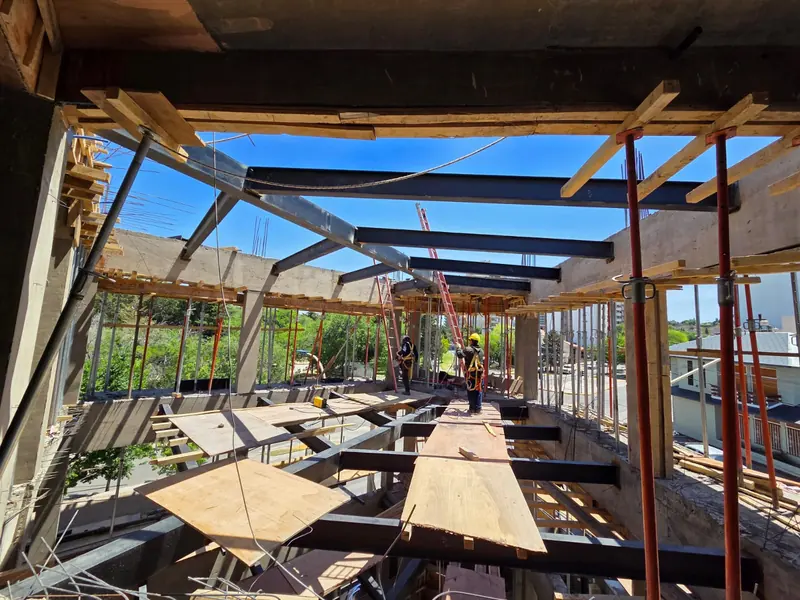 Floor level at the structural stage, with steel beams, formwork boards and props, and workers at the far end