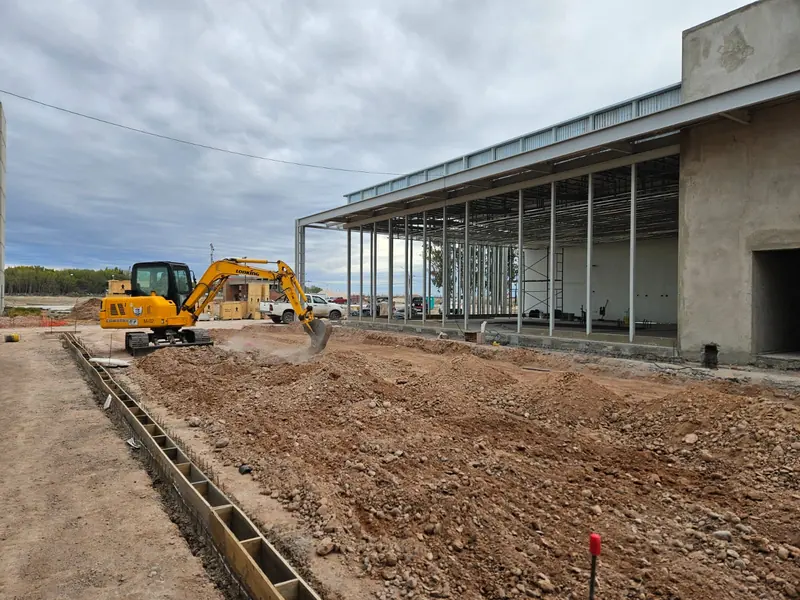 Excavator working next to the steel structure and concrete walls of the shop